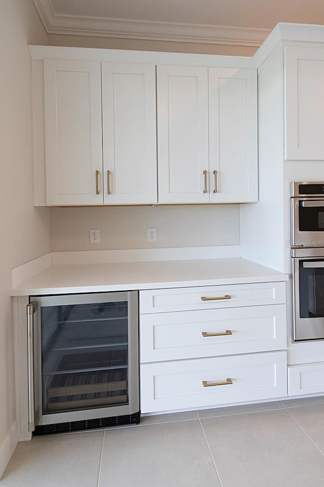 bar with stainless steel double oven, beverage cooler, crown molding, light tile patterned floors, and white cabinetry