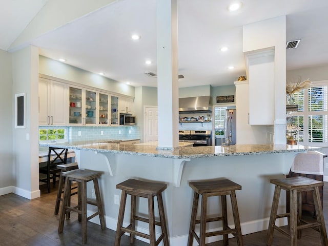 kitchen featuring white cabinetry, stainless steel appliances, wall chimney range hood, a kitchen breakfast bar, and kitchen peninsula