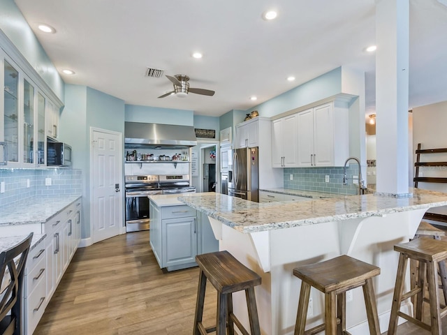 kitchen with white cabinets, appliances with stainless steel finishes, kitchen peninsula, and wall chimney exhaust hood