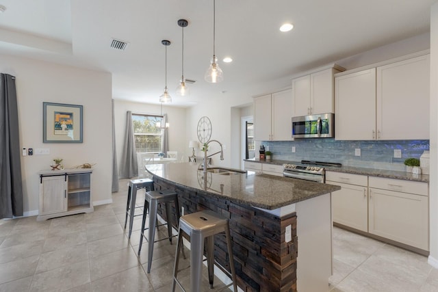 kitchen with white cabinets, appliances with stainless steel finishes, dark stone countertops, sink, and a center island with sink