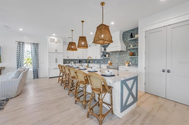 kitchen featuring custom exhaust hood, a breakfast bar, a large island with sink, white cabinets, and decorative light fixtures