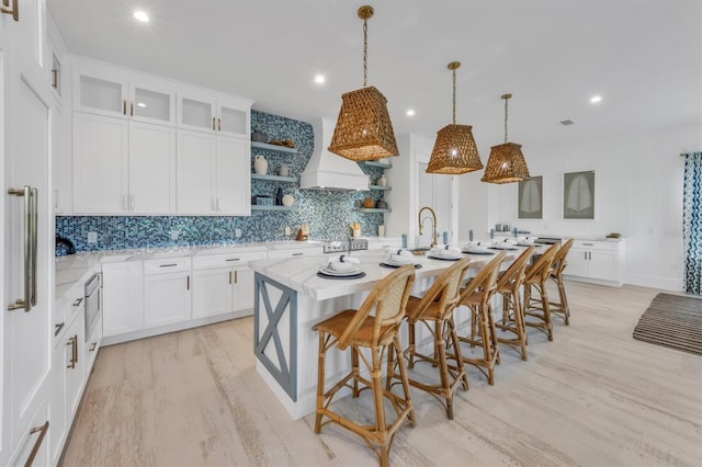 kitchen with a center island with sink, hanging light fixtures, custom range hood, light stone counters, and white cabinetry