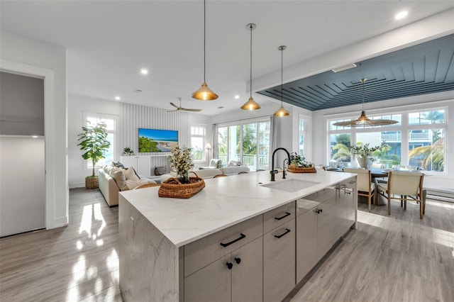 kitchen featuring a kitchen island with sink, sink, light hardwood / wood-style flooring, decorative light fixtures, and light stone counters