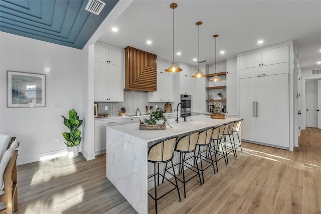 kitchen with a kitchen island with sink, hanging light fixtures, light hardwood / wood-style floors, decorative backsplash, and white cabinets