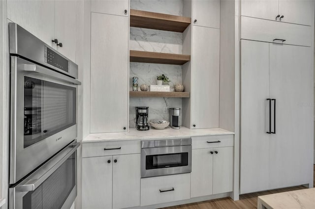 kitchen with light stone countertops, backsplash, stainless steel double oven, light hardwood / wood-style flooring, and white cabinetry