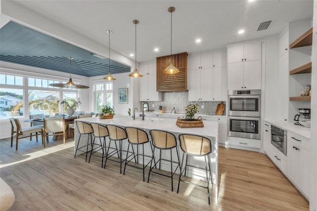 kitchen featuring white cabinets, light hardwood / wood-style flooring, an island with sink, double oven, and decorative light fixtures