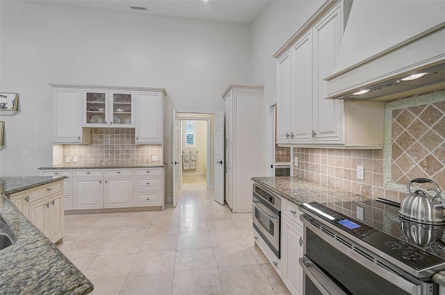 kitchen with a high ceiling, appliances with stainless steel finishes, custom exhaust hood, white cabinetry, and dark stone countertops