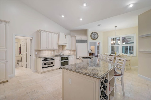 kitchen featuring lofted ceiling, premium range hood, oven, stainless steel oven, and a center island with sink