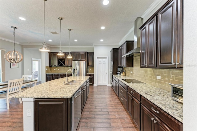 kitchen with appliances with stainless steel finishes, wall chimney exhaust hood, crown molding, a center island with sink, and hanging light fixtures