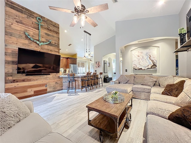 living room featuring arched walkways, ceiling fan, wooden walls, high vaulted ceiling, and light wood-type flooring