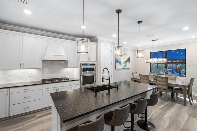 kitchen with premium range hood, white cabinets, stainless steel appliances, and an island with sink