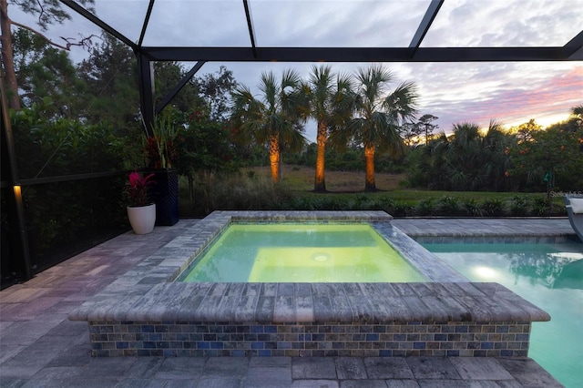 pool at dusk featuring a patio, glass enclosure, and an in ground hot tub