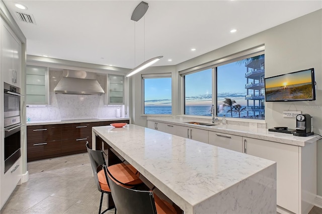 kitchen featuring wall chimney exhaust hood, a center island, dark brown cabinets, pendant lighting, and white cabinets