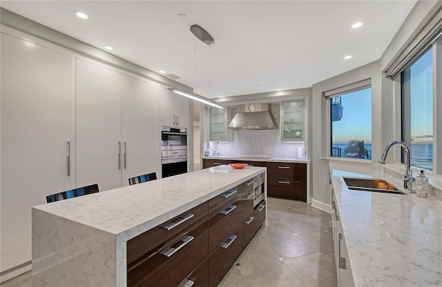 kitchen with wall chimney range hood, sink, white cabinets, and a kitchen island