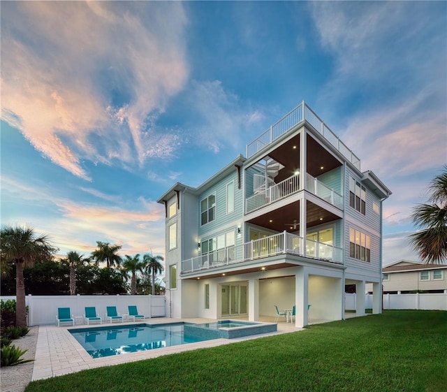 back house at dusk featuring a lawn, a patio area, a balcony, a pool with hot tub, and ceiling fan