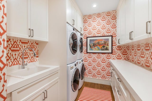 clothes washing area featuring wood finished floors, wallpapered walls, a sink, cabinet space, and stacked washing maching and dryer