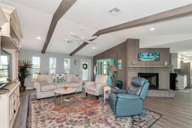 living room featuring wood walls, vaulted ceiling with beams, a tiled fireplace, ceiling fan, and light wood-type flooring