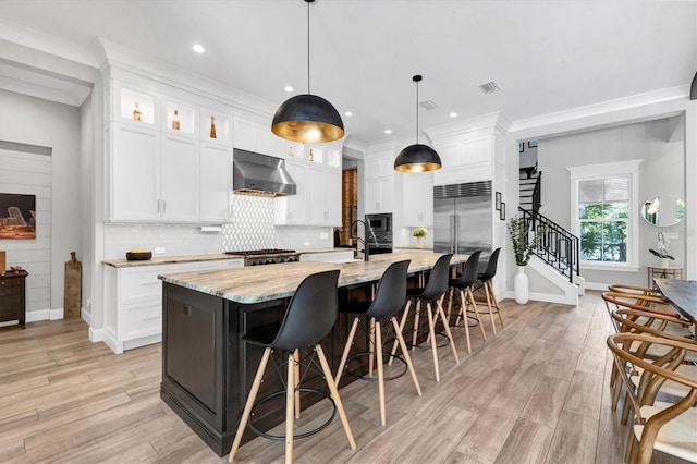 kitchen with light stone counters, white cabinetry, an island with sink, and range hood