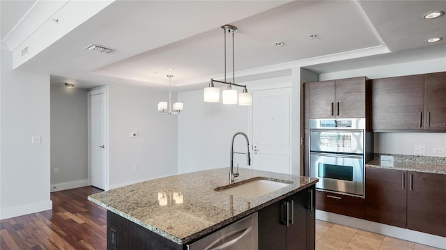 kitchen with sink, stainless steel appliances, light stone counters, an island with sink, and a raised ceiling