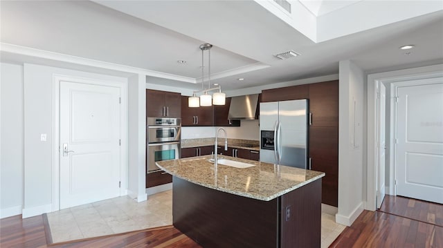 kitchen with sink, dark brown cabinets, a center island with sink, stainless steel appliances, and wall chimney range hood