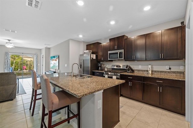 kitchen with light stone counters, a center island with sink, a kitchen bar, appliances with stainless steel finishes, and ceiling fan