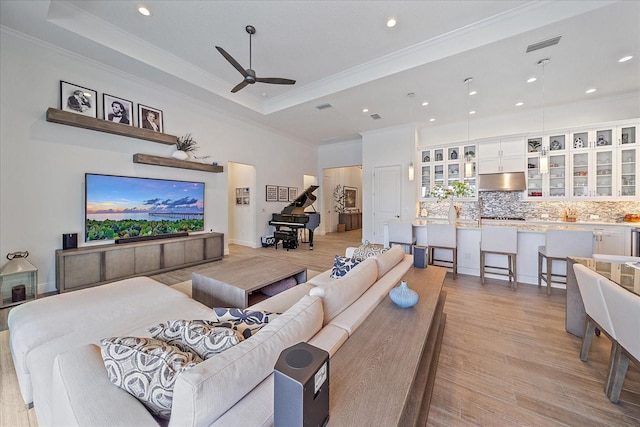 living room with a raised ceiling, visible vents, ornamental molding, light wood-style floors, and a ceiling fan