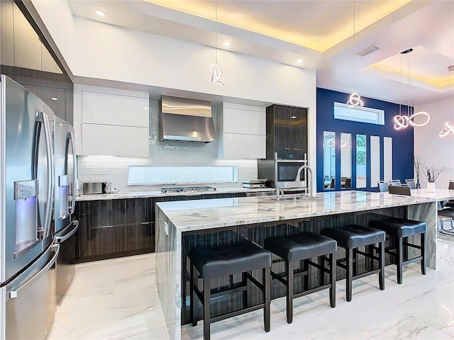 kitchen with pendant lighting, wall chimney range hood, stainless steel appliances, and an island with sink