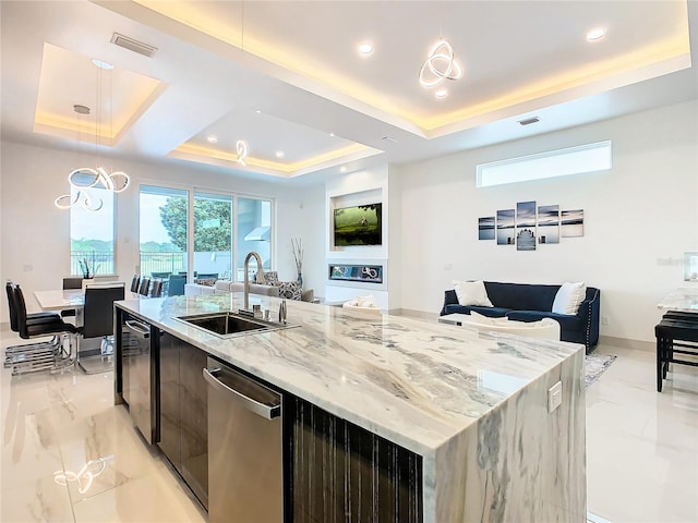 kitchen with light stone counters, a tray ceiling, sink, a center island with sink, and dishwasher