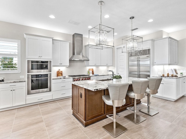 kitchen with wall chimney exhaust hood, an island with sink, appliances with stainless steel finishes, white cabinetry, and a breakfast bar area