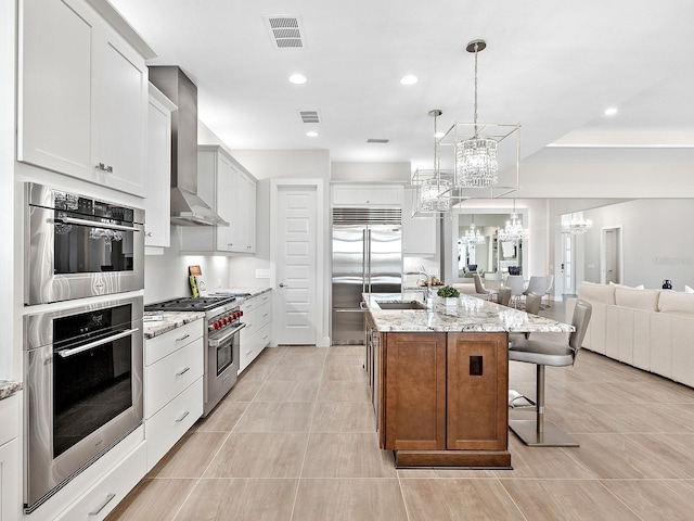 kitchen featuring light stone countertops, wall chimney exhaust hood, a kitchen island with sink, high quality appliances, and white cabinetry