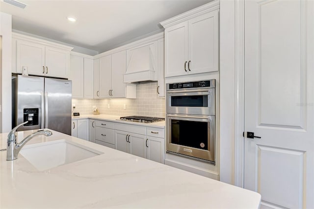 kitchen featuring tasteful backsplash, appliances with stainless steel finishes, white cabinets, a sink, and premium range hood