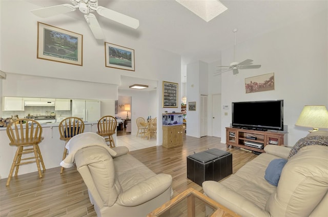 living room featuring ceiling fan, a skylight, high vaulted ceiling, and light hardwood / wood-style flooring