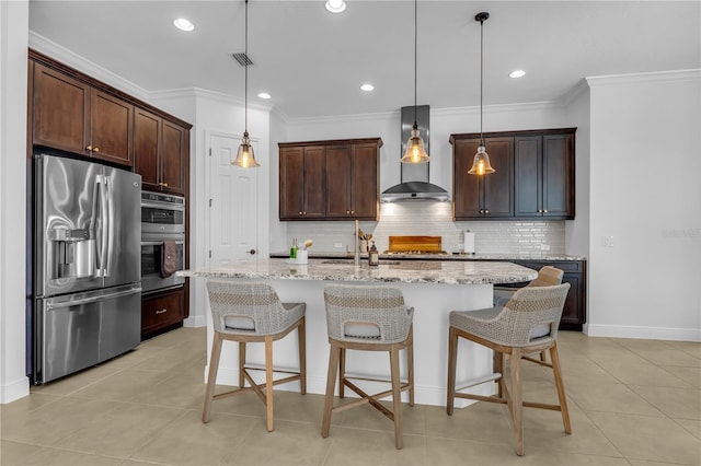 kitchen featuring pendant lighting, wall chimney exhaust hood, light stone countertops, an island with sink, and appliances with stainless steel finishes