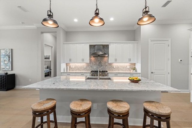 kitchen featuring decorative light fixtures, white cabinets, an island with sink, and wall chimney range hood