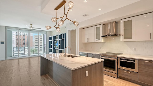 kitchen with wall chimney range hood, sink, electric range, an island with sink, and white cabinetry