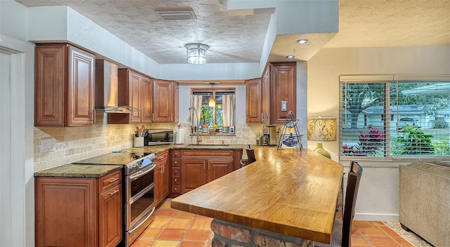 kitchen with stainless steel appliances, a sink, decorative backsplash, wall chimney exhaust hood, and brown cabinetry