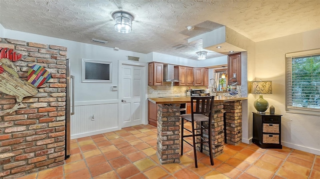 kitchen with brown cabinets, visible vents, wainscoting, wall chimney range hood, and a textured ceiling