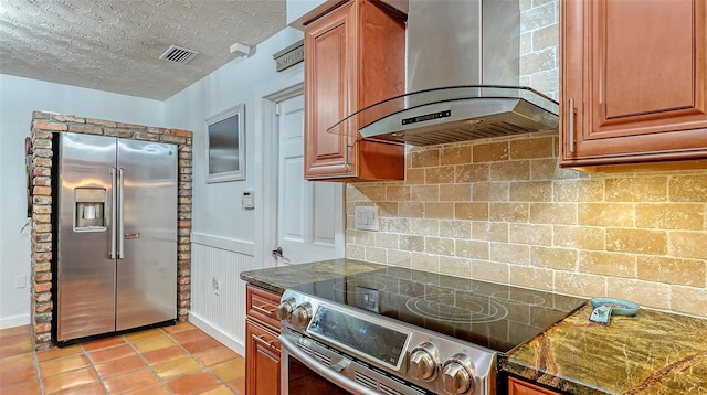 kitchen with a textured ceiling, stainless steel appliances, visible vents, wall chimney range hood, and brown cabinets