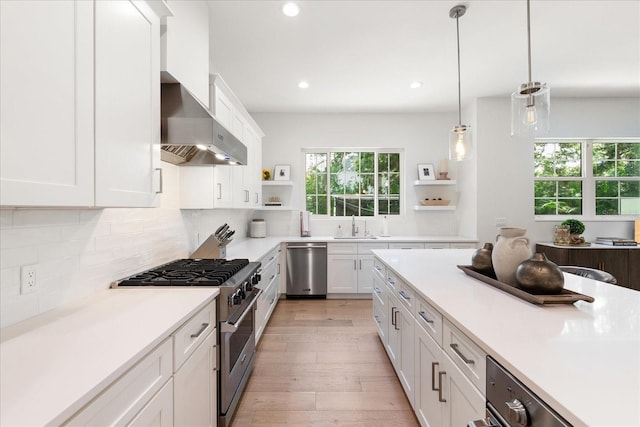 kitchen featuring wall chimney range hood, light hardwood / wood-style floors, decorative light fixtures, white cabinets, and appliances with stainless steel finishes