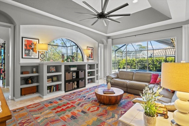 tiled living room with ceiling fan, built in features, and a tray ceiling