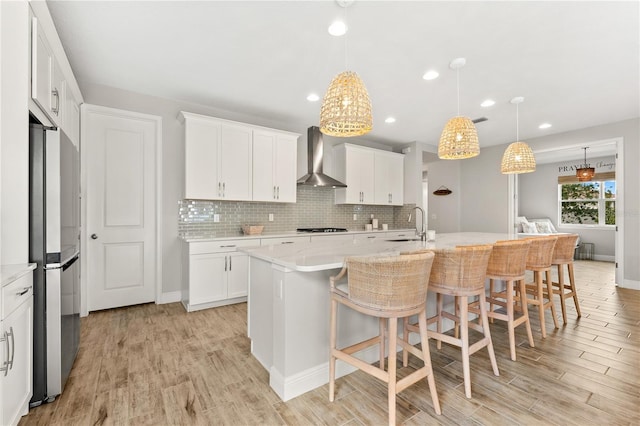 kitchen featuring a kitchen island with sink, wall chimney range hood, stainless steel fridge, decorative light fixtures, and white cabinetry