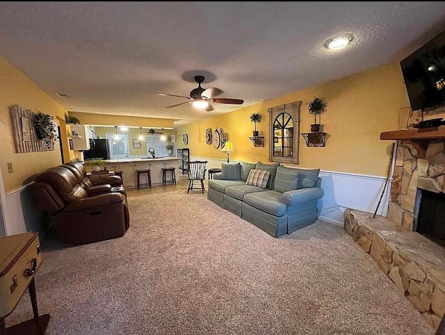 carpeted living room featuring ceiling fan, a stone fireplace, and a textured ceiling