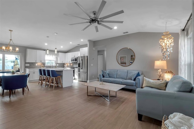 living room with ceiling fan with notable chandelier, light wood-type flooring, sink, and vaulted ceiling