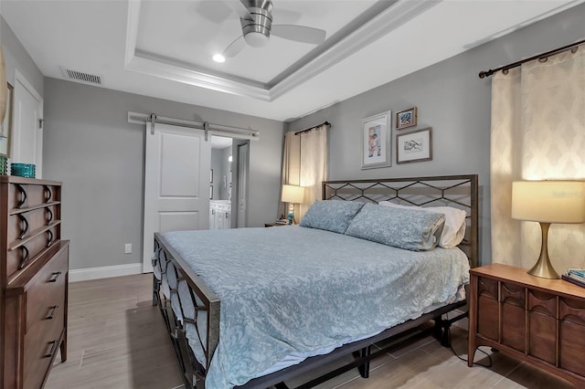 bedroom featuring ensuite bath, ceiling fan, a barn door, a tray ceiling, and wood-type flooring