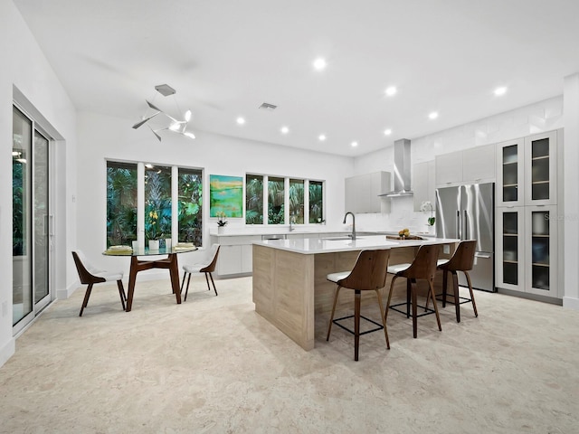 kitchen with gray cabinetry, a kitchen breakfast bar, a large island with sink, stainless steel built in fridge, and wall chimney exhaust hood