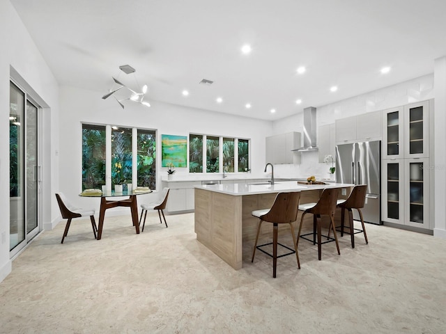 kitchen with a large island with sink, stainless steel fridge, modern cabinets, wall chimney exhaust hood, and visible vents