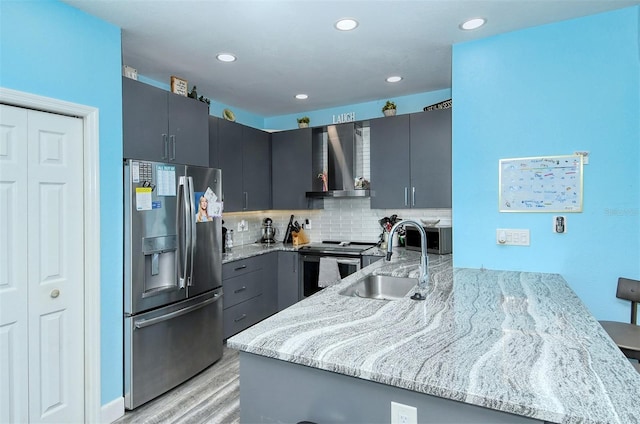 kitchen with wall chimney range hood, sink, appliances with stainless steel finishes, light stone counters, and kitchen peninsula