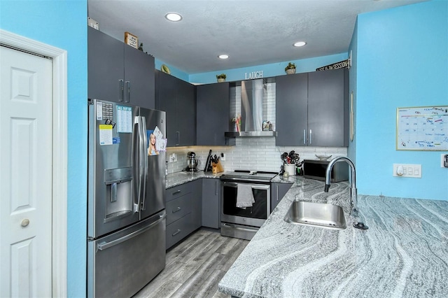 kitchen featuring wall chimney exhaust hood, light stone counters, sink, and appliances with stainless steel finishes