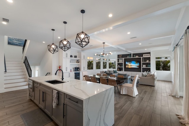 kitchen featuring a kitchen island with sink, hanging light fixtures, beam ceiling, light stone counters, and sink