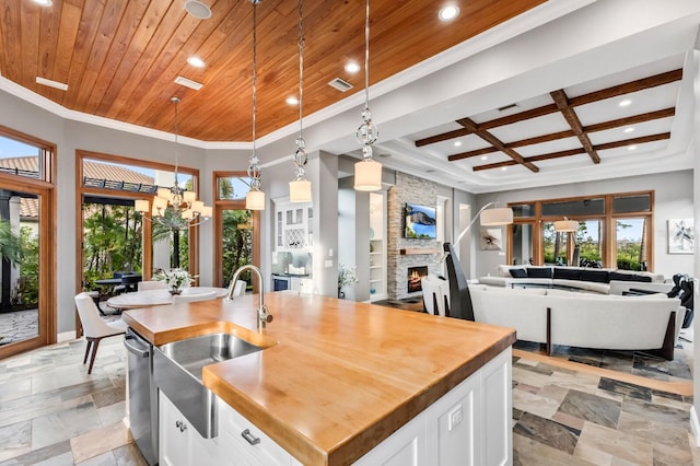 kitchen with coffered ceiling, white cabinetry, hanging light fixtures, sink, and an island with sink